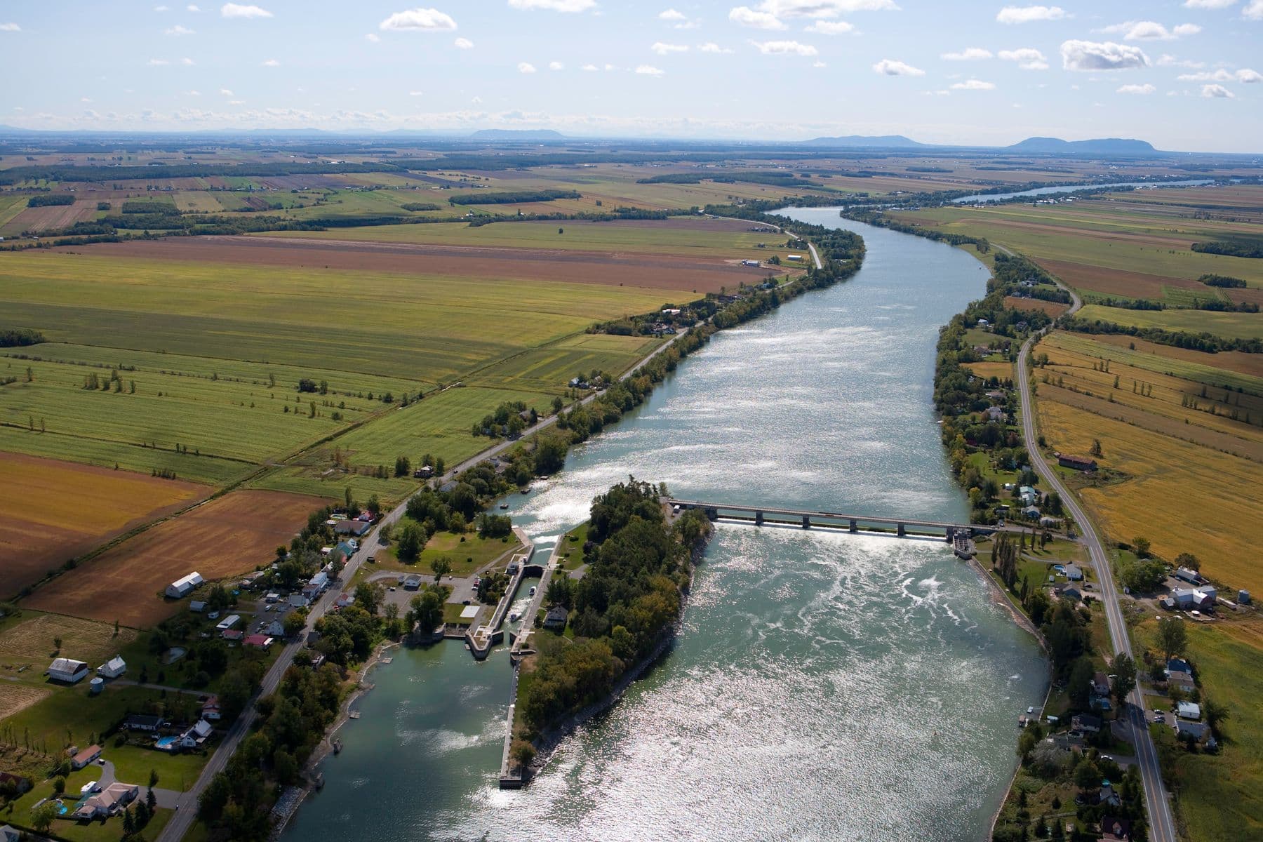 Vue aérienne d’un canal traversant une campagne agricole avec écluse, routes sinueuses et champs colorés à perte de vue.