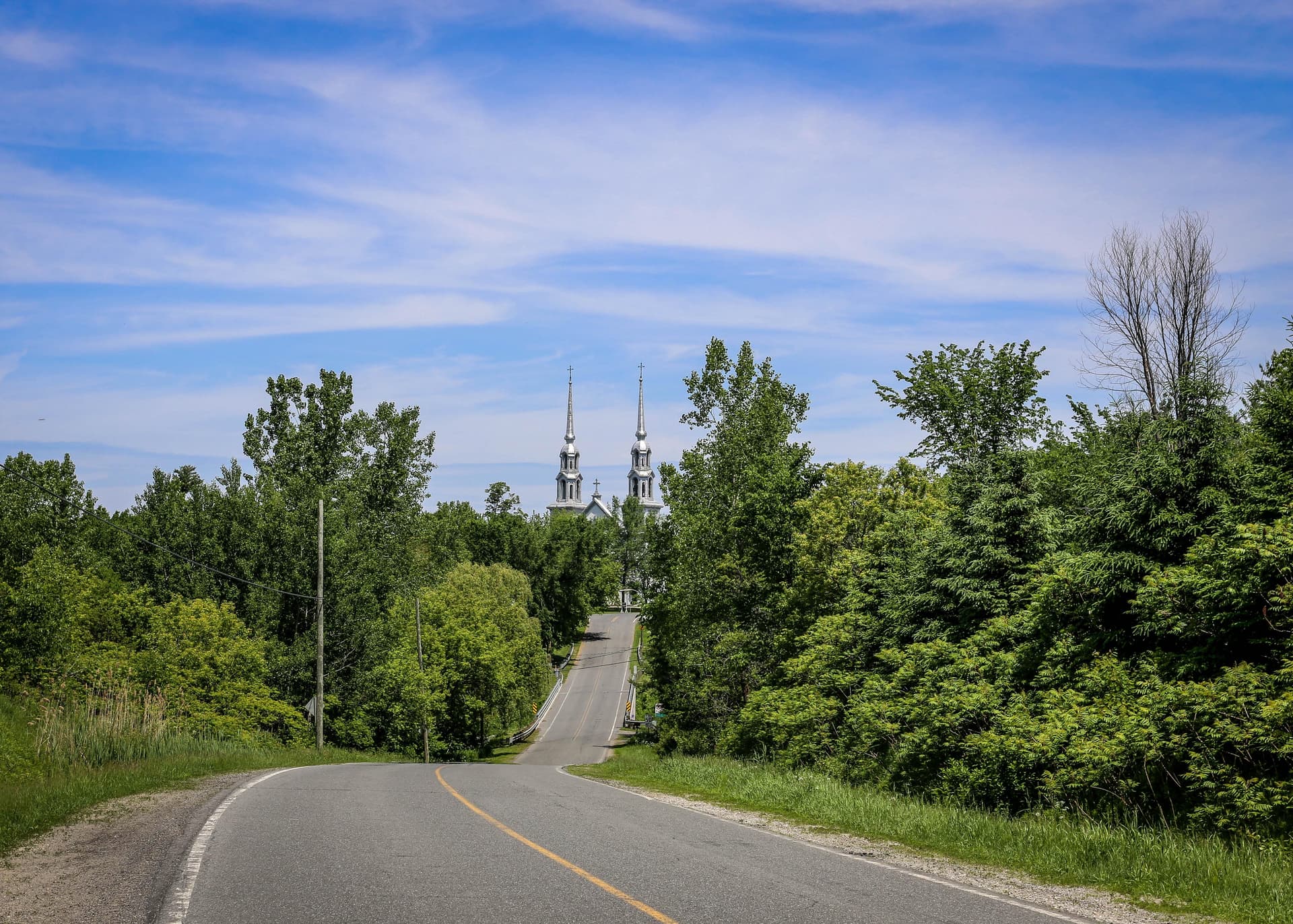 Route sinueuse bordée d’arbres menant à une église aux clochers argentés visibles à l’horizon sous un ciel bleu.
