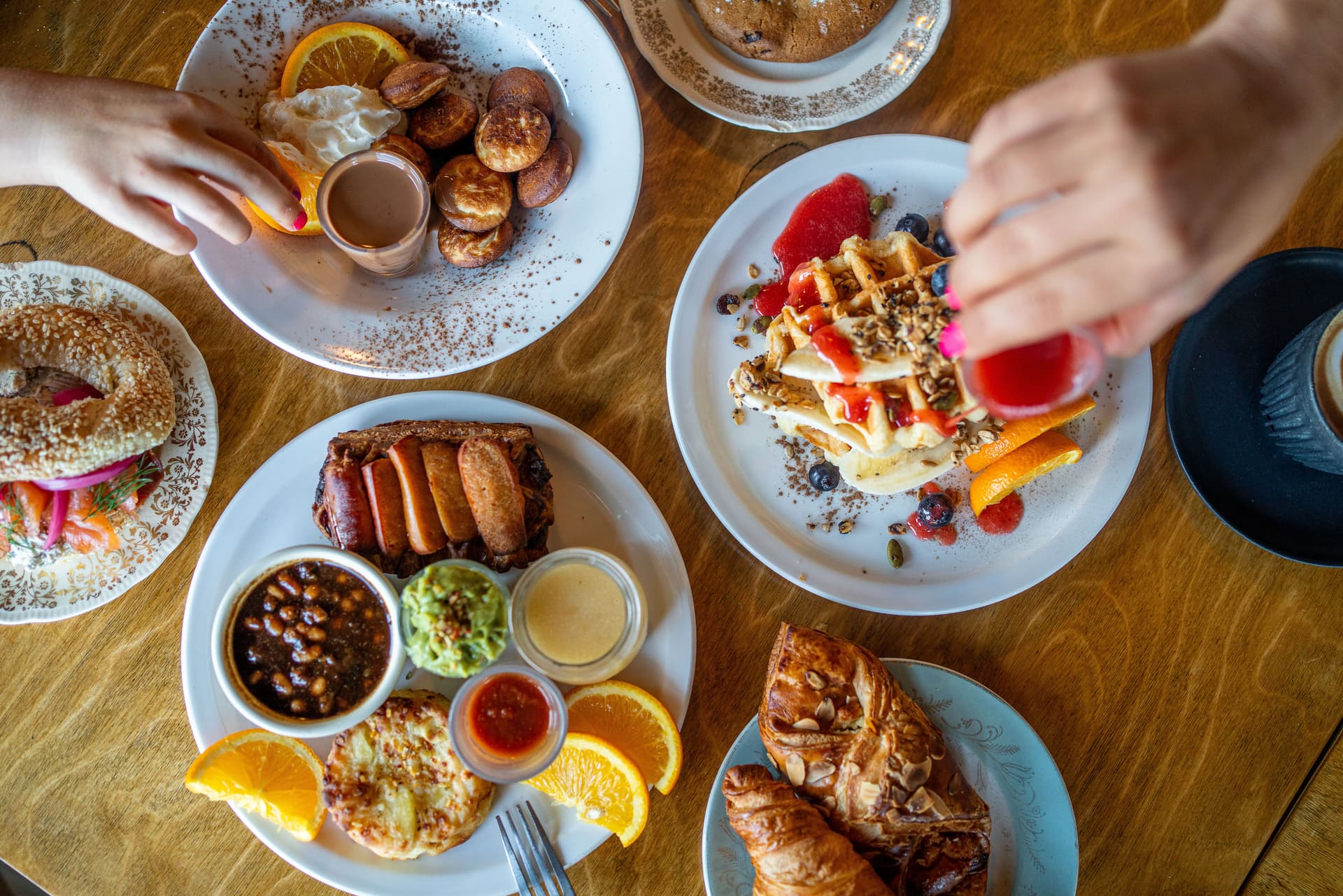 Table de brunch garnie de gaufres aux fruits, croissants, bagel, saucisses, sauces variées et mains en train de se servir.