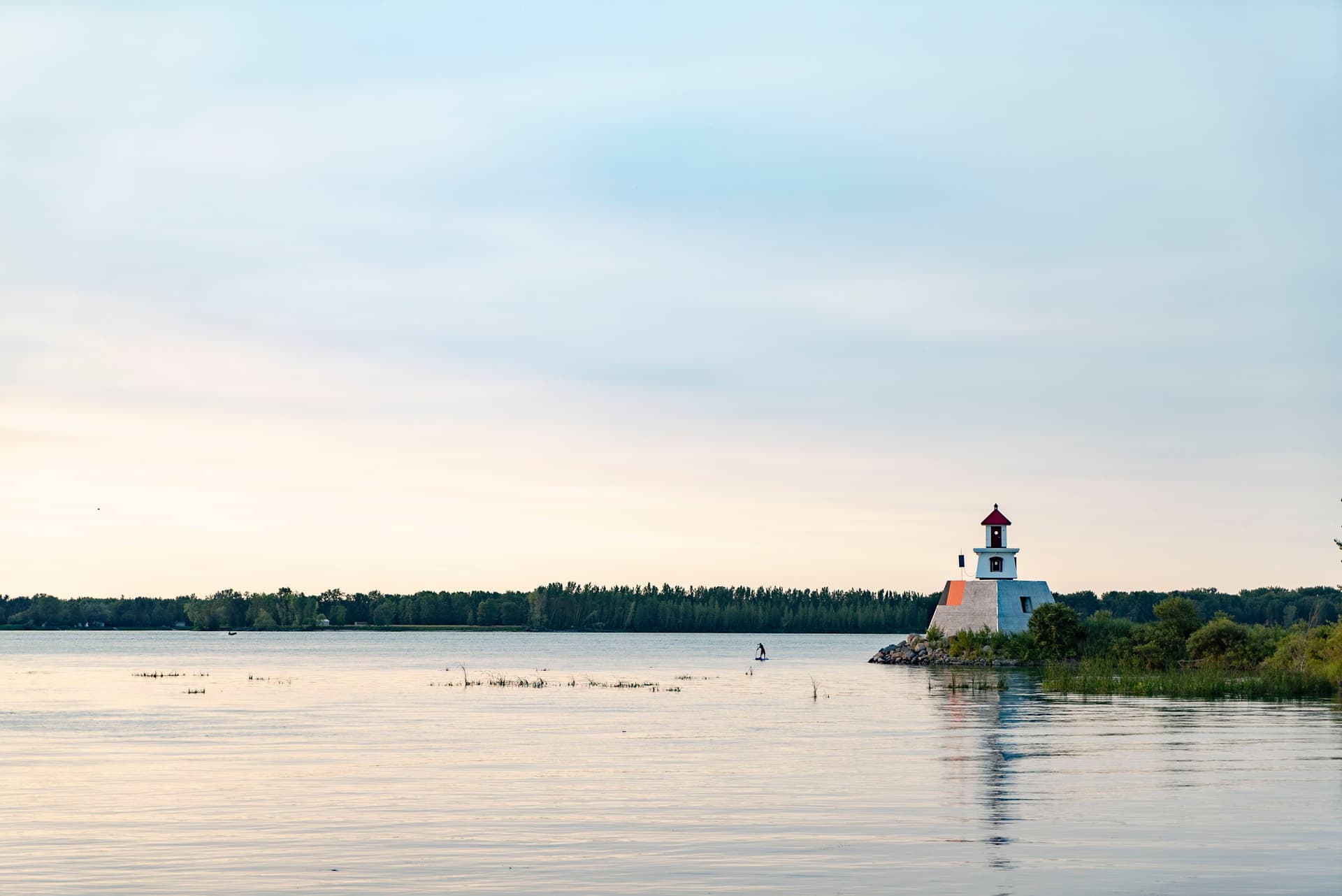 Petit phare blanc à toit rouge au bord d’un lac calme, entouré de verdure sous un ciel pastel.