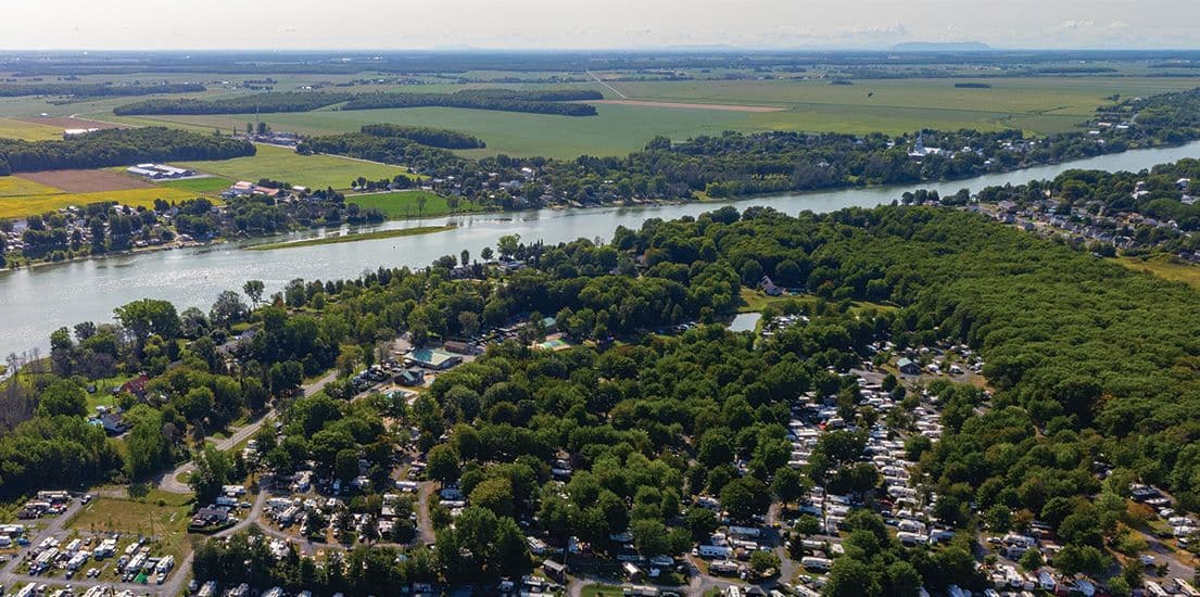 Vue aérienne d’un vaste terrain de camping boisé longeant une rivière sinueuse, avec champs agricoles à l’horizon.