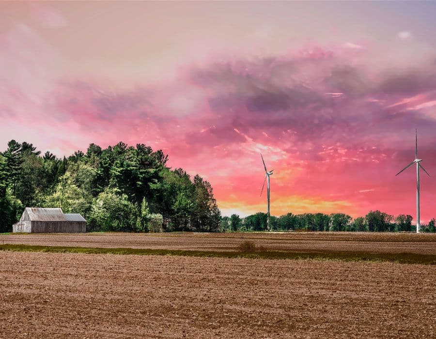 Champ labouré avec éoliennes et petite grange en bois, bordé d’arbres sous un ciel rose et violet au coucher du soleil.
